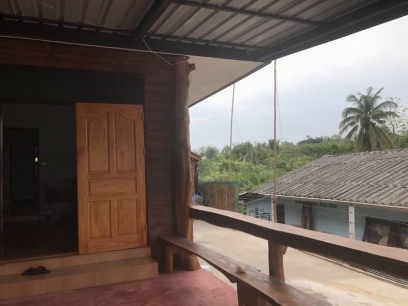 A wooden entrance door is showcased, leading into the homestay. A spacious wooden bench runs alongside the wall, with a view of lush greenery and distant rooftops in the background. The area is shaded, providing a relaxing outdoor space.