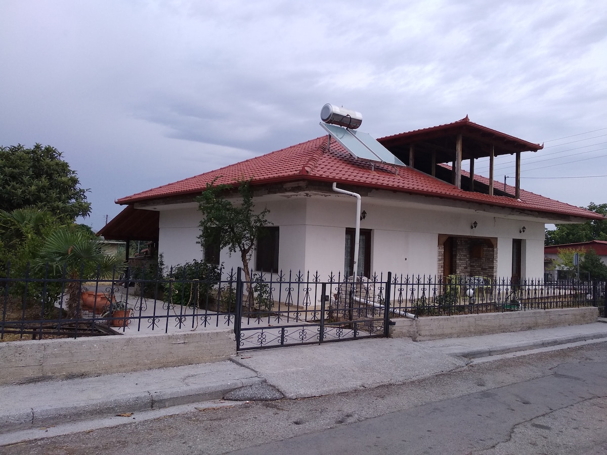 A single-story villa is displayed with a red-tiled roof and a prominent solar panel. The exterior features a spacious fenced yard with a variety of greenery, providing shade and a welcoming entryway. A covered terrace extends from the main building.