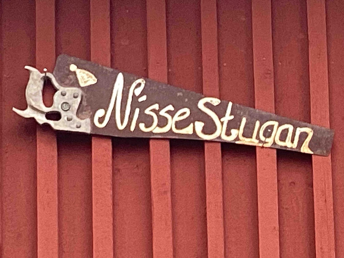 A wooden sign is displayed on the exterior of the red cottage, featuring the name 'Nisse Stugan' in an artistic script. The sign is mounted on a textured wooden surface, showcasing traditional craftsmanship.