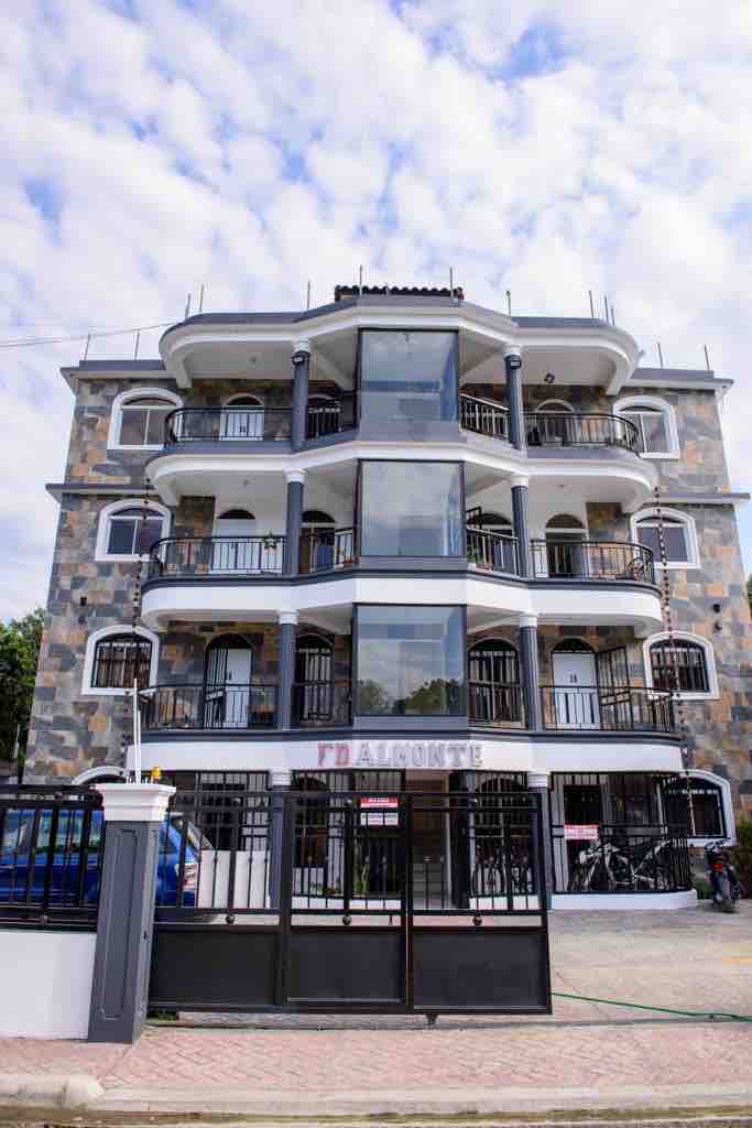 The exterior of the building features a modern design with multiple levels, large glass windows, and decorative stone accents. Balconies with wrought iron railings enhance the façade, while the entrance is framed by a gated entryway. A clear blue sky complements the structure.