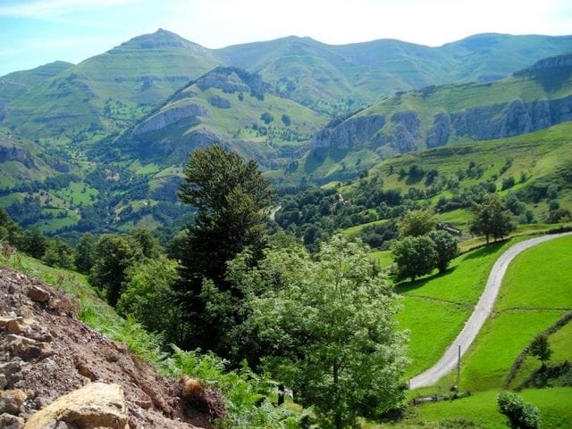 Rolling green hills and majestic mountains are visible in the background, creating a serene landscape. Lush trees and open fields are interspersed with winding roads, inviting exploration of the natural surroundings.