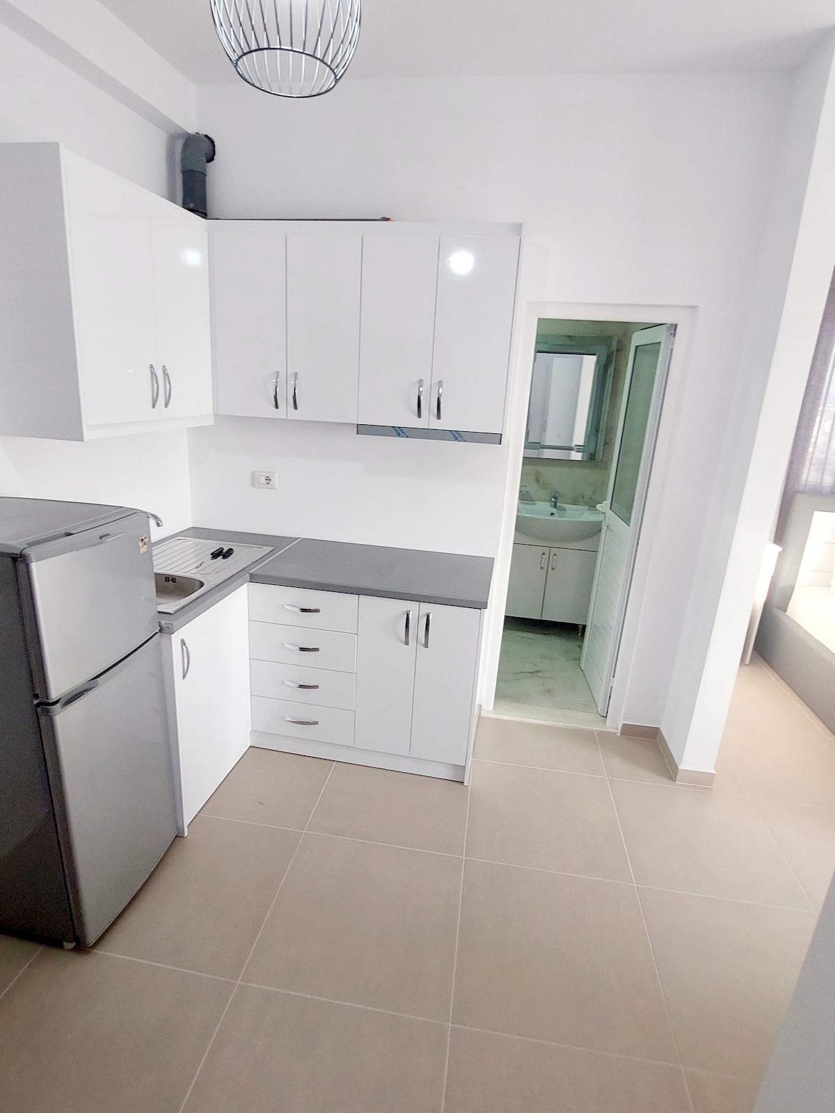A modern kitchen is displayed, featuring sleek white cabinets and stainless steel appliances. The refrigerator stands prominently beside the countertop, which is equipped with a stove. An open doorway leads to a bathroom, while light enters from a nearby window.
