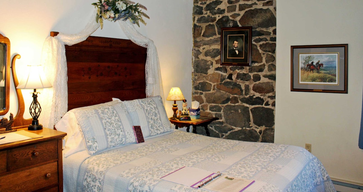 A cozy sleeping area features a queen bed with a patterned quilt, flanked by a wooden nightstand and a classic lamp. The exposed stone wall adds character, enhanced by framed artwork and a decorative mirror above a rustic dresser.