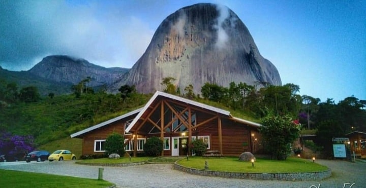 A wooden lodge is set against a large, rocky mountain partially shrouded in clouds. The structure features large windows and a steeply sloped roof, surrounded by landscaped greenery. A gravel path and parking area are visible in the foreground.