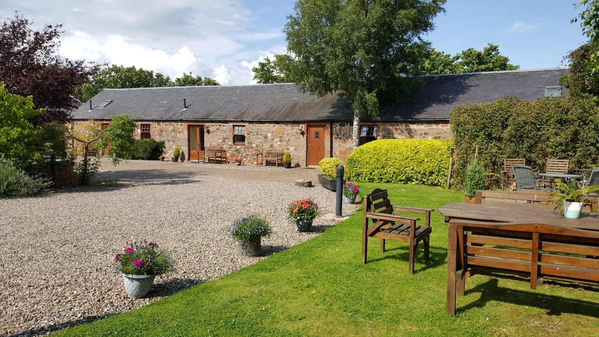 A charming courtyard view showcases stone stable block apartments surrounded by well-maintained gardens. Gravel pathways meander through lush greenery and vibrant flowerpots. Wooden seating arrangements are positioned on the lawn, offering a peaceful space for relaxation amidst the rural atmosphere.