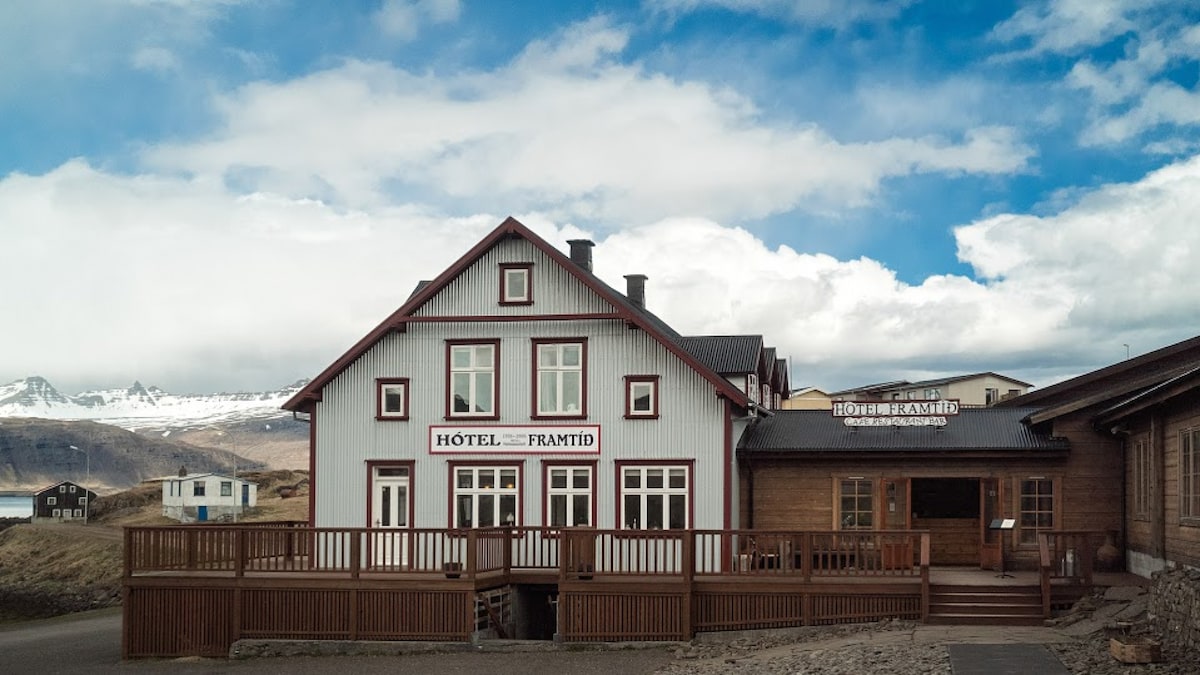 The exterior of Hótel Framtíð features a two-story building with a wooden deck, large windows, and traditional architectural details. Snow-capped mountains can be seen in the background against a partly cloudy sky.