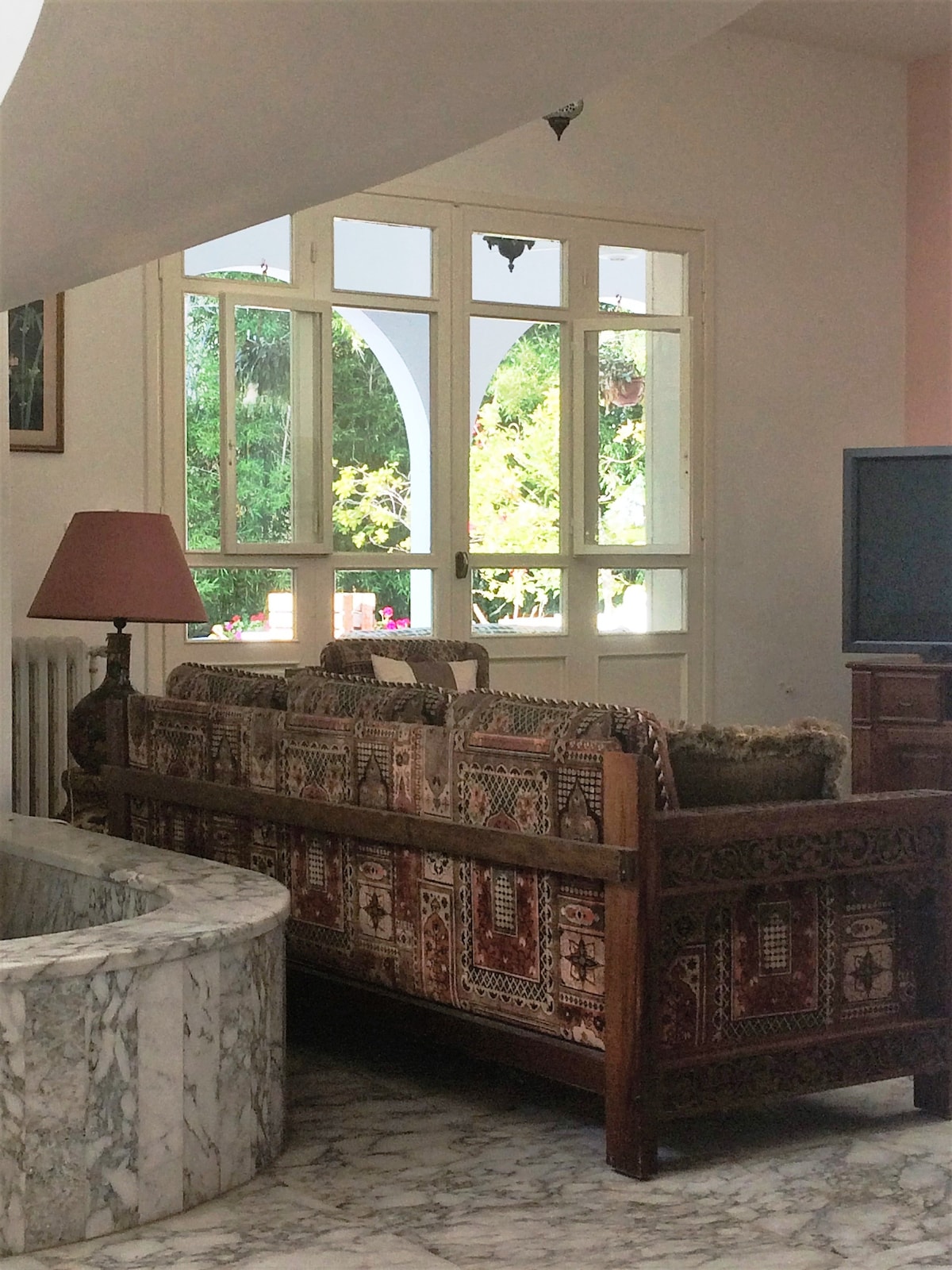 A traditional wooden sofa with intricate carvings is positioned near a large window, allowing natural light to fill the space. A decorative lamp stands on a side table next to the sofa, while the marble flooring adds elegance to the room.