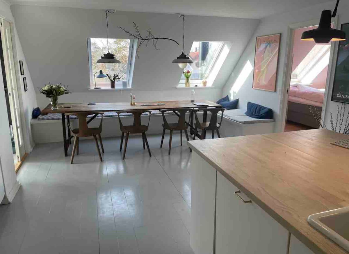 A bright kitchen and dining area features a long wooden table surrounded by eight chairs. Two skylights flood the space with natural light, highlighting the simple, modern décor. A cozy seating nook is visible by the window, with a glimpse of the adjacent bedroom in the background.