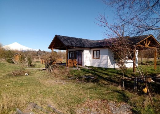A single-story house is showcased with a sloped roof and a front porch. Lush green grass surrounds the home, with trees and plants enriching the landscape. Snow-capped mountains are visible in the background, highlighting the natural beauty of the setting.