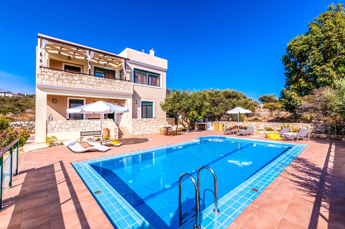 A modern villa with a private swimming pool sits on a sunlit terrace. Sun loungers and umbrellas provide relaxation options beside the pool, while lush greenery frames the area. The facade features a stone-clad design, complemented by spacious balconies overlooking the scenic surroundings.