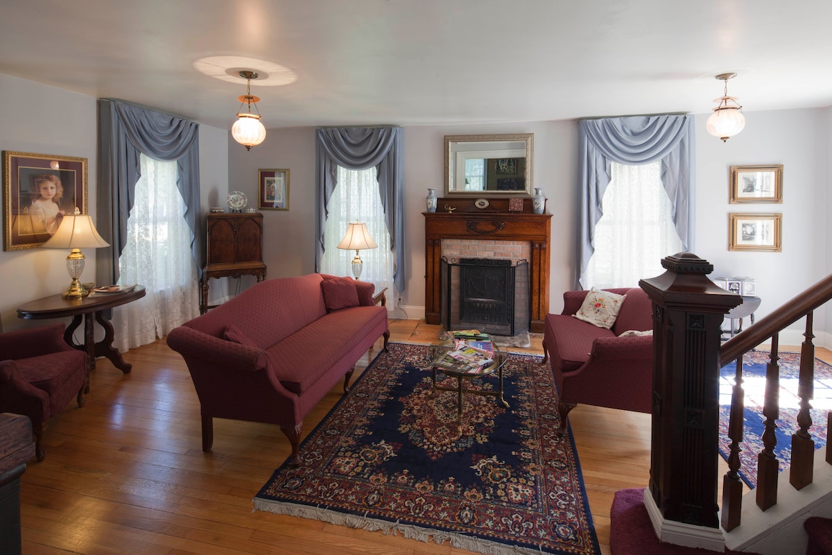 A welcoming living room features a fireplace surrounded by elegant furnishings. Two red sofas sit across from each other, with a wooden table in between. Soft lighting from lamps complements window drapes. A decorative area rug adds warmth to the hardwood floor.