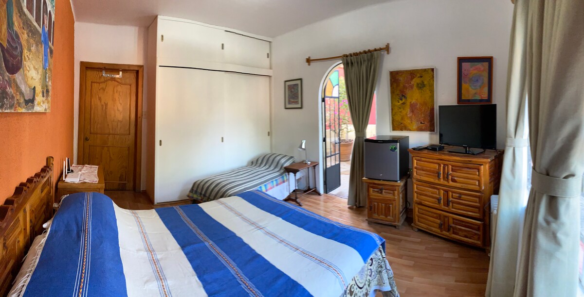 A spacious room is showcased, featuring a large bed with a blue-striped blanket and a sofa bed along the side. Wooden furniture, including a dresser and a TV stand, complements the decor. Natural light is filtered through curtains, illuminating artwork on the walls.
