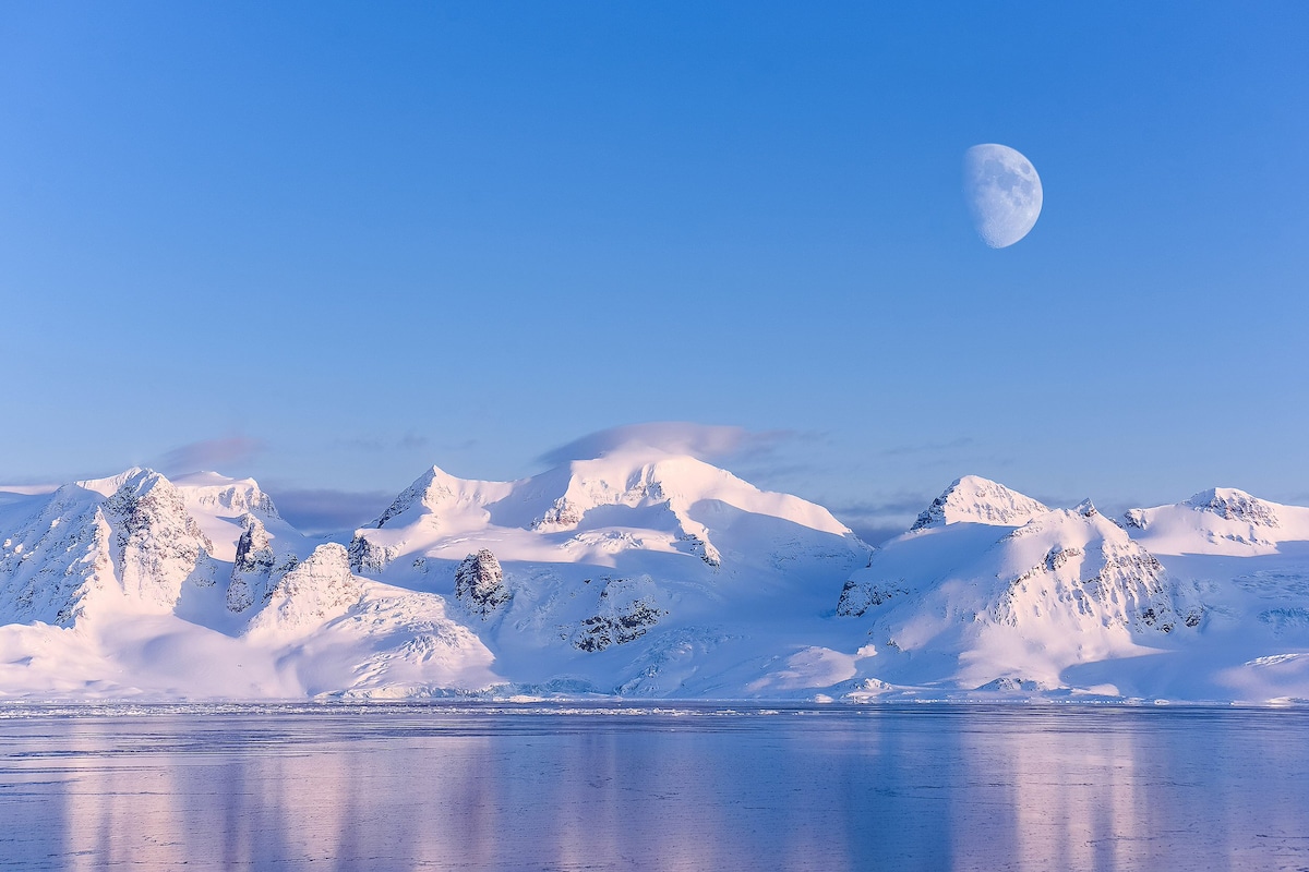 A vast landscape features snow-covered mountains reflecting in a tranquil body of water, illuminated by soft hues of blue and white. A large moon hangs over the scene, enhancing the serene atmosphere of the arctic environment.
