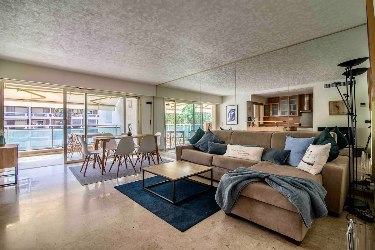 A spacious living area features a large beige sofa facing a coffee table on a navy blue rug. Mirrored walls reflect natural light from sliding glass doors that open to a balcony with outdoor seating. A dining area with white chairs is visible in the background.