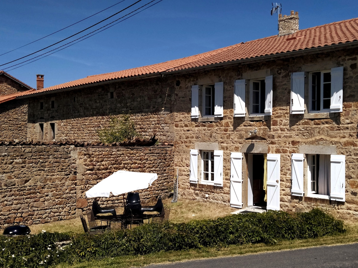 A charming stone house with white shutters is set against a clear blue sky. A covered outdoor seating area is visible, featuring a table and six chairs. Lush greenery frames the property, enhancing the inviting outdoor space.