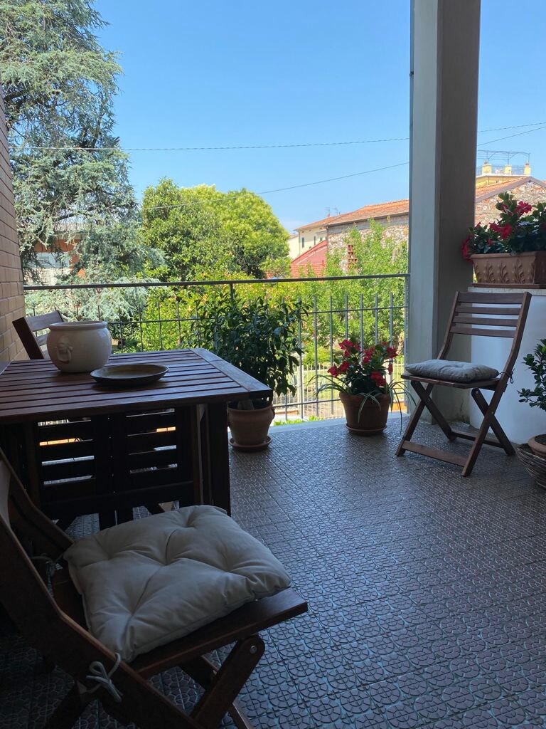 An outdoor terrace is presented with a wooden table and two chairs, surrounded by potted plants. The area offers a view of greenery and nearby buildings under a clear blue sky, providing a peaceful space for relaxation.