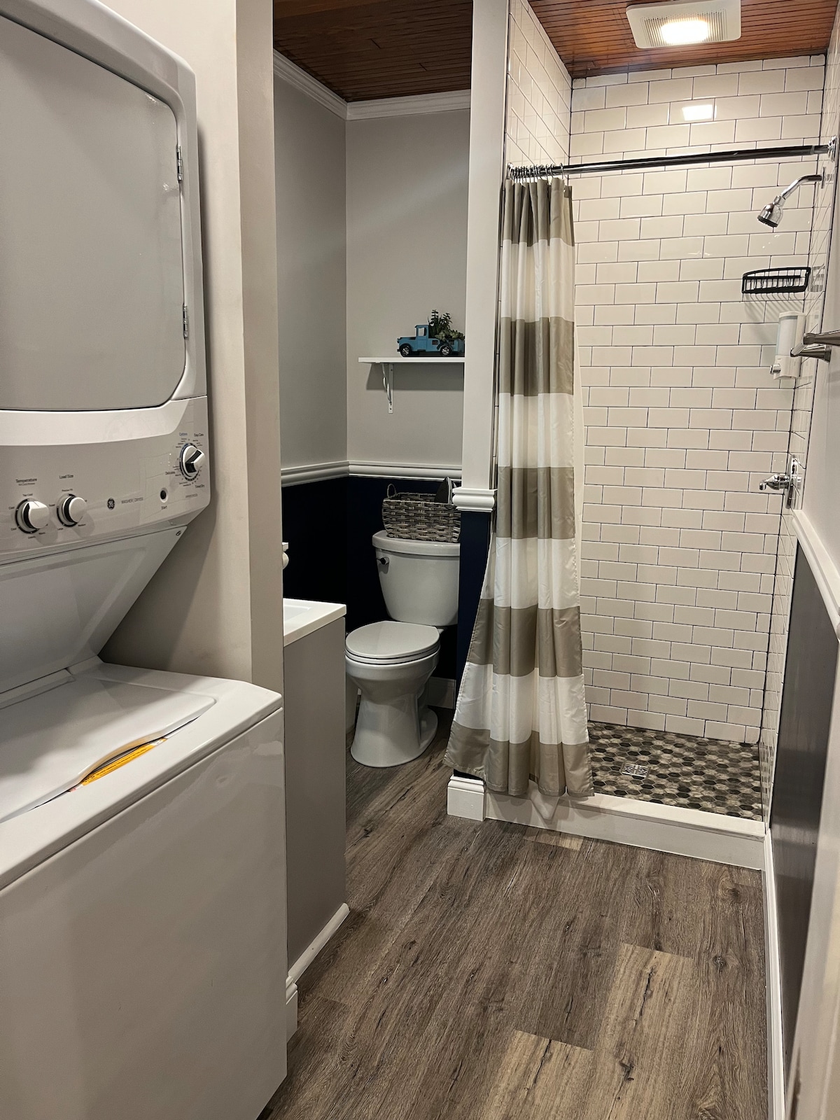 The bathroom area features a modern shower with a white tiled wall and decorative pebble flooring. A washing machine and dryer are available, positioned next to a toilet. A striped shower curtain adds a soft touch to the space, which is complemented by warm wood accents.