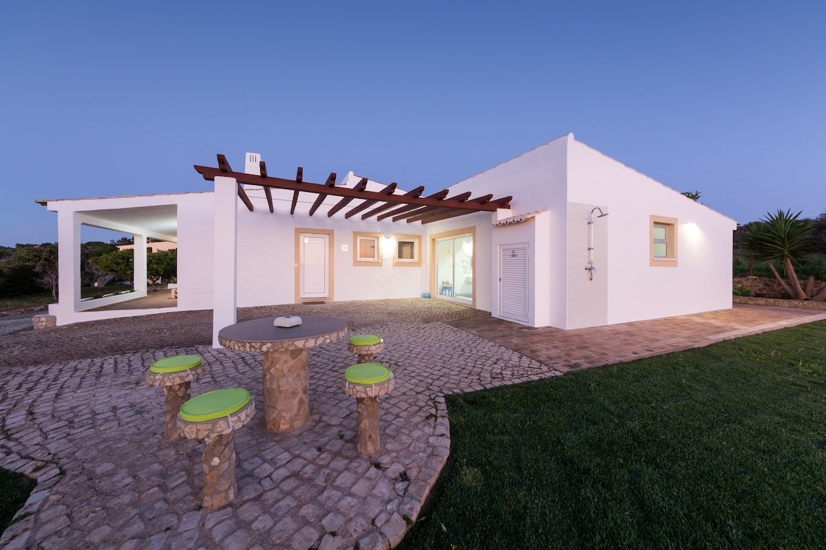 A modern white house features a spacious outdoor area with stone seating and bright green accents. An outdoor shower is positioned nearby, and the cobblestone path leads to an inviting entrance. Lush greenery accentuates the tranquil surroundings, with large windows allowing natural light to enter.