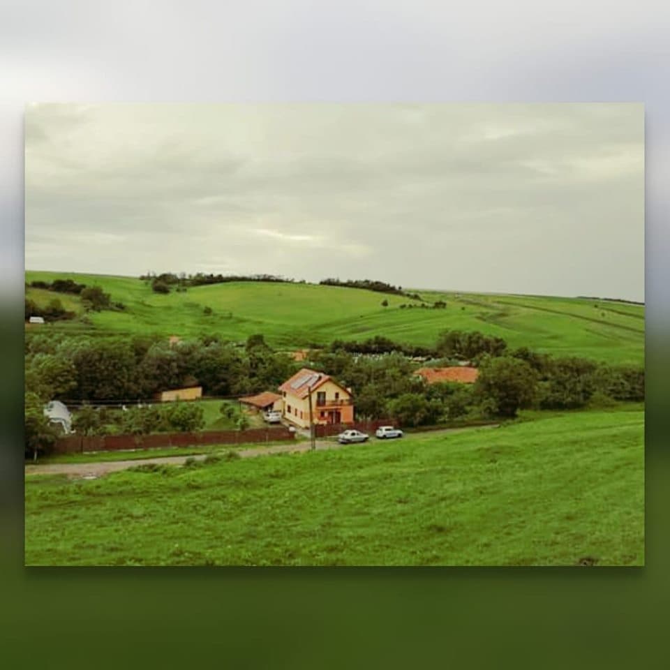 A scenic view of Pensiunea Casa Murani is presented, surrounded by lush green hills. The building features a distinct yellow facade and is nestled among greenery, with visible cars parked along a gravel road in the foreground, highlighting the natural landscape.
