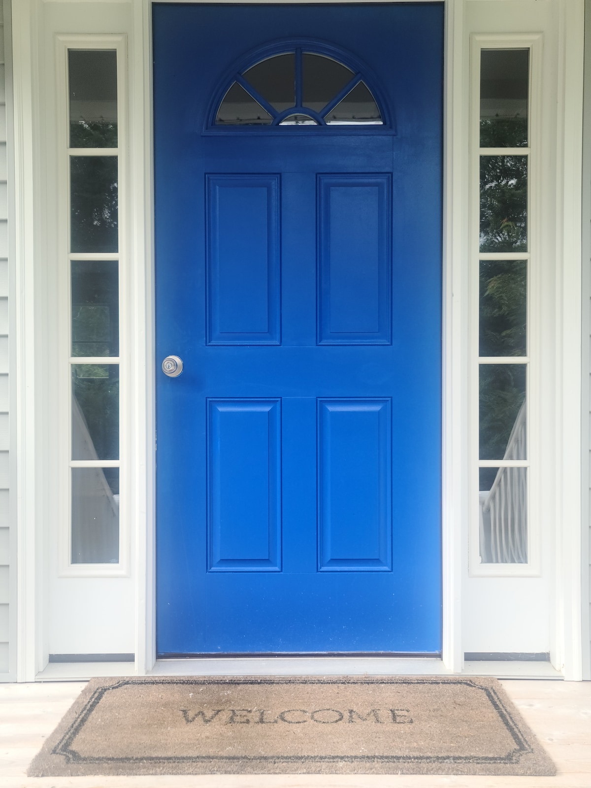 A vibrant blue front door is framed by two tall, narrow windows on either side. The entrance is completed with a welcome mat positioned centrally in front of the door, providing a clear, inviting access to the home.