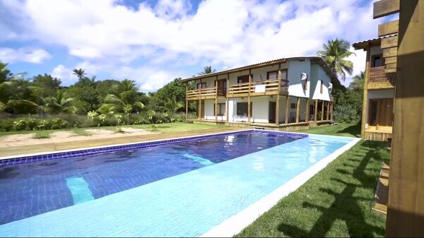 The image features a well-maintained outdoor pool, bordered by lush greenery. A two-story building with balconies is positioned alongside the pool, while a clear sky and soft clouds add to the tranquil setting. The overall ambiance suggests a relaxing retreat.