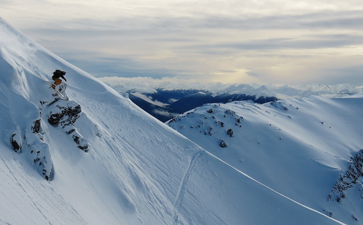 A vast snow-covered landscape unfolds, showcasing rolling hills and distant mountain peaks. Soft clouds hover above, complemented by a subdued sky that transitions from light to dark. A single skier glides gracefully down a slope, leaving a faint trail behind in the untouched snow.