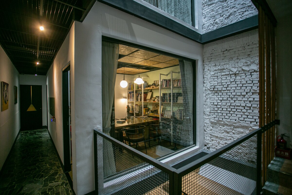 A well-lit common area is visible through a large window, featuring a table surrounded by chairs and bookshelves filled with neatly organized books. Natural light flows into the space, highlighting the textures of the walls and ceiling.