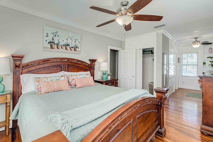 The master bedroom features sturdy furniture, ample natural light, a dressing room leading to the bathroom, two ceiling fans, and a large TV on the dresser with cable and Roku streaming. It’s a comfortable and functional space for relaxation.