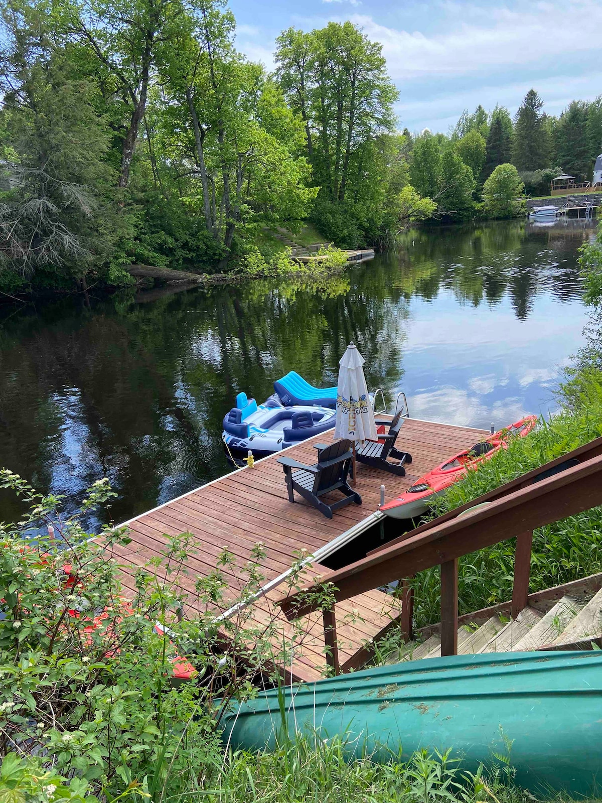 A wooden dock extends into a calm river, surrounded by lush greenery. A cozy seating area includes two chairs and an umbrella, while colorful kayaks are positioned nearby. A large inflatable float rests on the water, reflecting the clear blue sky.