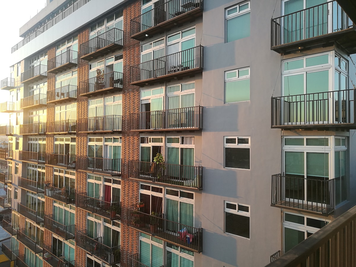 The modern building features multiple floors with numerous large windows and private balconies. Warm evening light reflects off the glass, enhancing the attractive façade. A variety of plants can be seen on some balconies, adding subtle greenery to the structure.