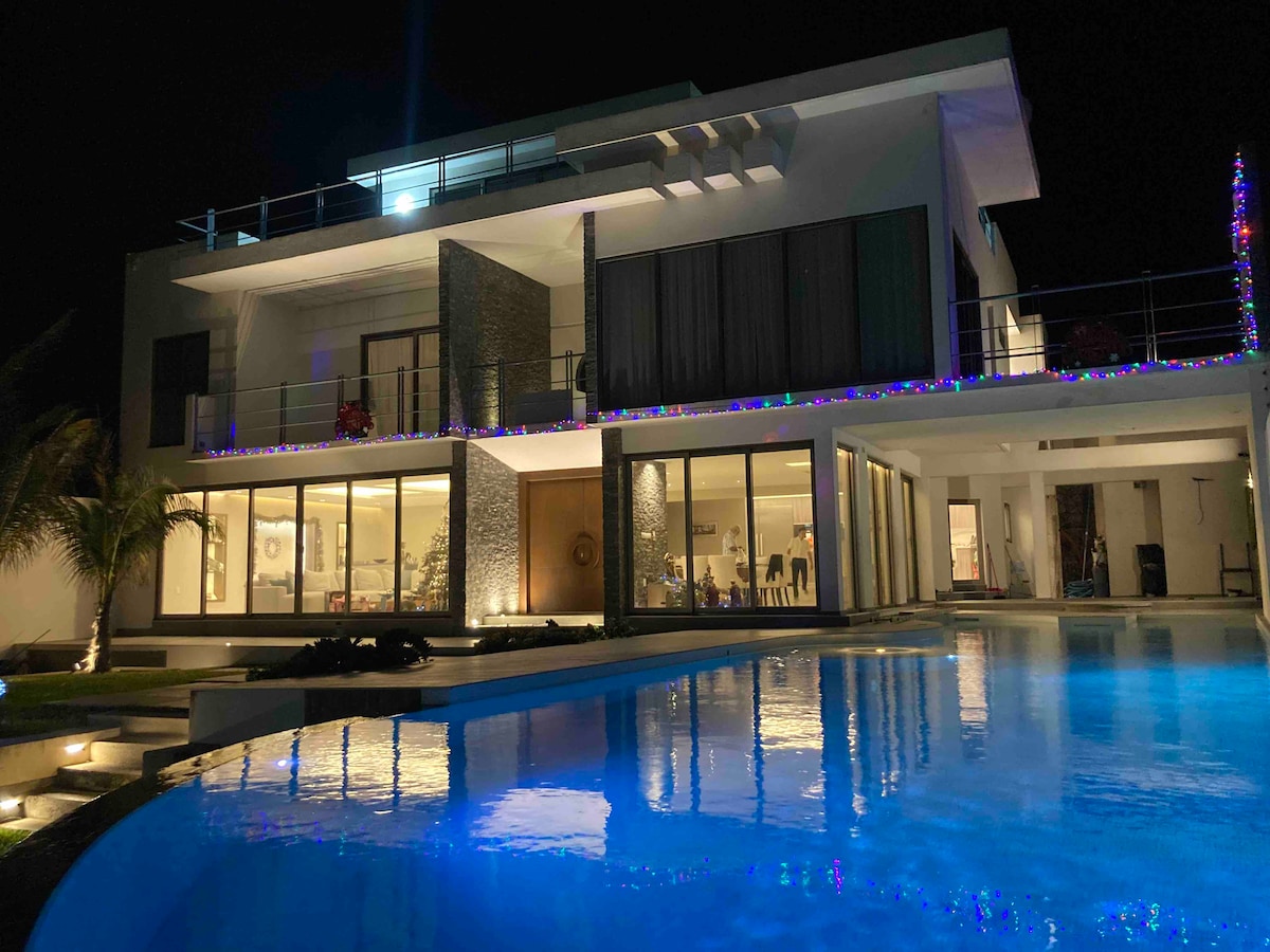 A modern three-story villa is illuminated at night, showcasing expansive glass windows that reflect the surrounding lights. A serene pool stretches out in front, complemented by manicured landscaping and palm trees. The structure features a spacious terrace, enhancing outdoor living experiences.