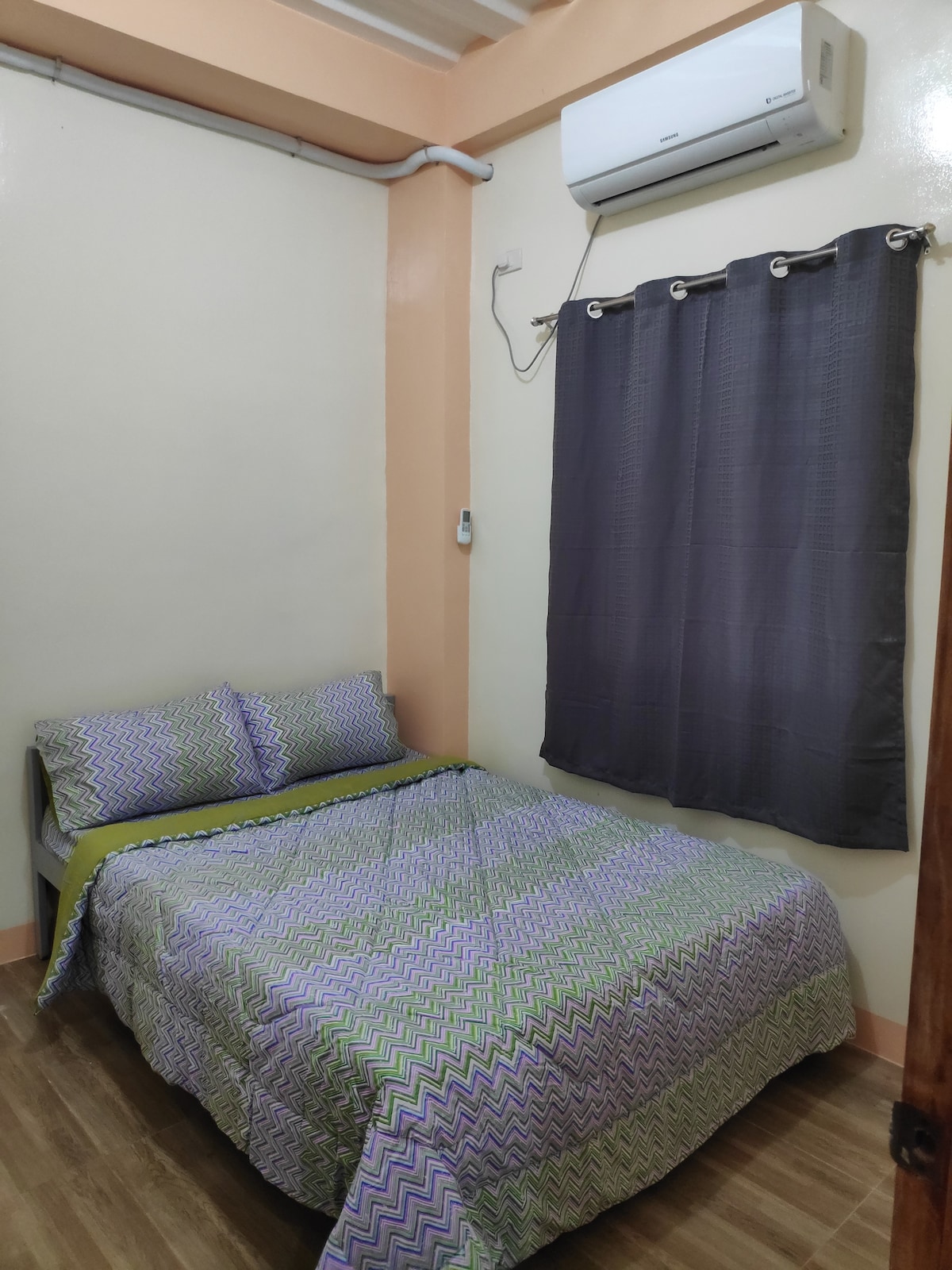 A twin bedroom features a spacious bed with a green and patterned blanket. The room is equipped with an air conditioning unit mounted above the bed. A dark curtain partially covers a window, while the floor is finished with light-colored wood.