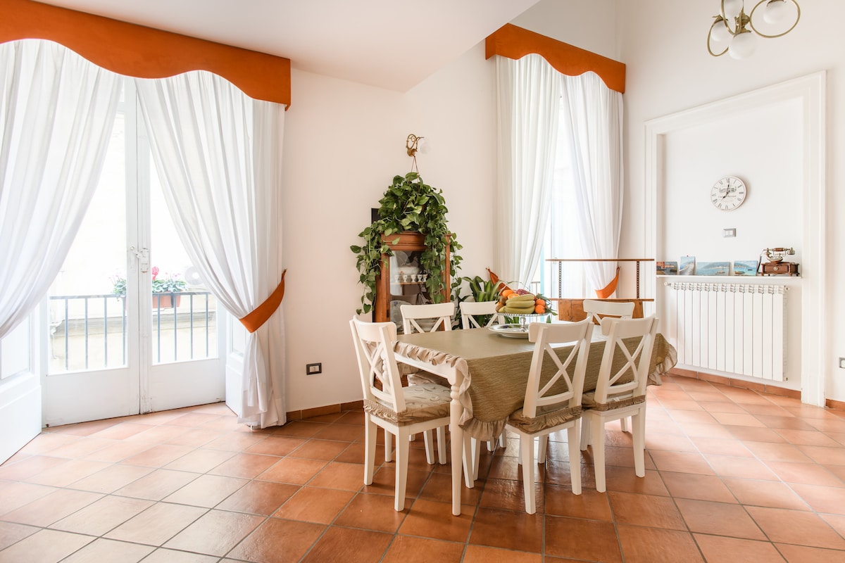 The dining area features a large table surrounded by white chairs. Natural light filters through large windows, adorned with light curtains. A green plant adds a touch of nature, while the warm terracotta floor complements the inviting atmosphere.