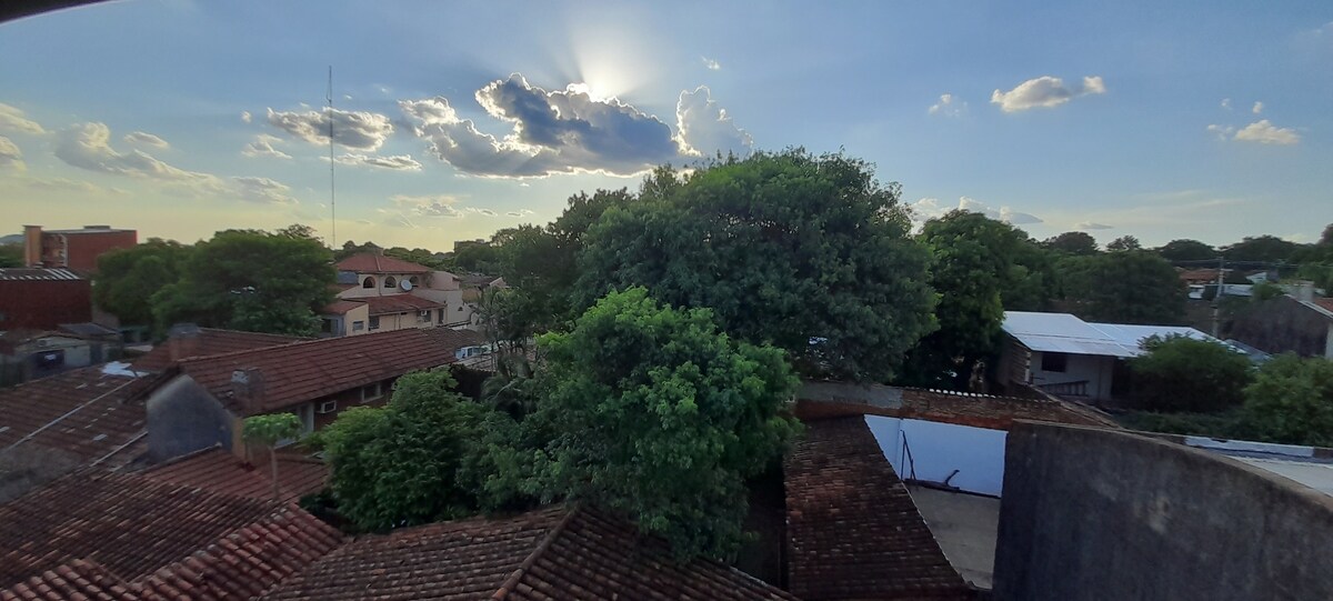 A scenic view of the neighborhood reveals lush green trees and terracotta rooftops. The sky is bright with scattered clouds, showcasing a sunset that softly illuminates the surrounding area. The peaceful residential environment is evident in the mixture of buildings and greenery.