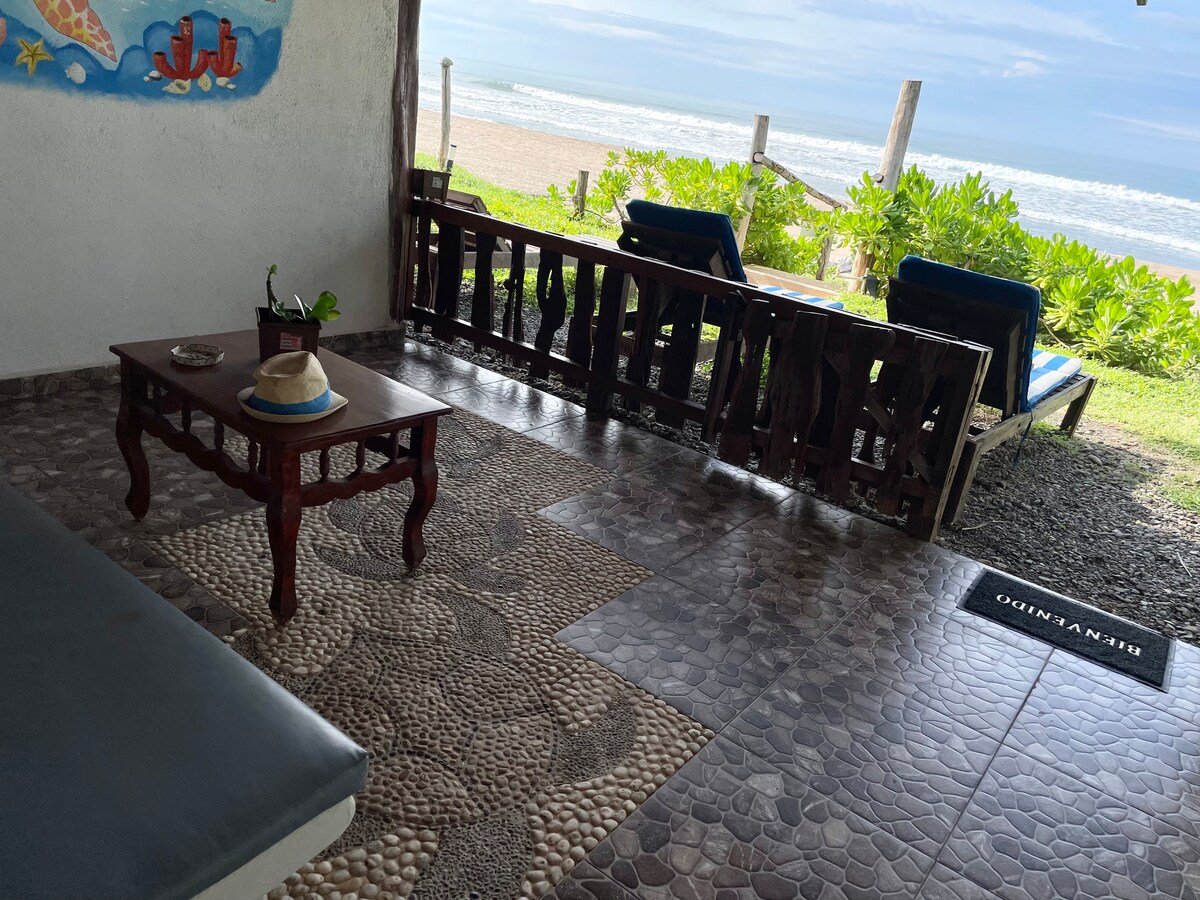 A shaded terrace area is visible, featuring a wooden table with a hat resting on it. Two lounge chairs face the beach, offering views of the ocean and surrounding greenery. The flooring is adorned with unique stone patterns, enhancing the outdoor space's natural vibe.
