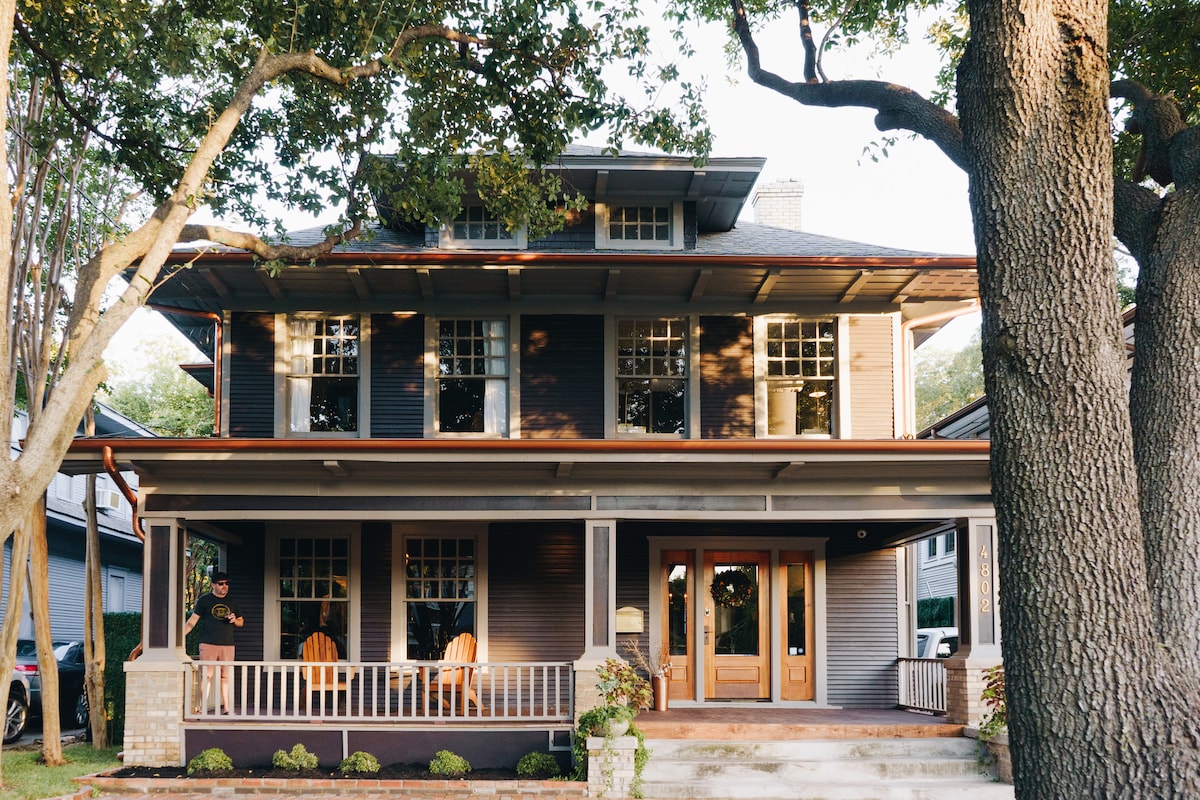 The exterior of a historic three-story house features large windows and a welcoming porch with multiple rocking chairs. The structure is shaded by mature trees, with natural landscaping enhancing the entrance. A wooden door stands prominently, inviting guests into the home.