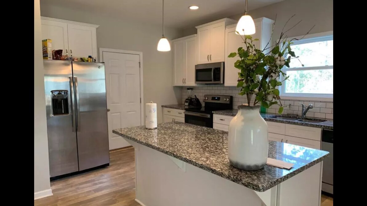 A contemporary kitchen is highlighted by a large granite island at its center, complemented by pendant lighting. Stainless steel appliances, including a refrigerator and microwave, are visible against white cabinetry. A vase with greenery adds a touch of nature to the inviting space.