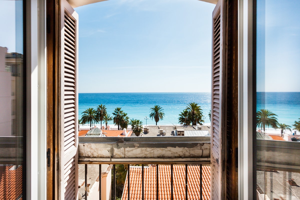 Two large shutters are open to reveal a stunning view of the sea and palm trees under a clear blue sky. The balcony railing is visible, framed by the warm tones of the apartment. Sunlight brightens the interior space, enhancing the serene atmosphere.