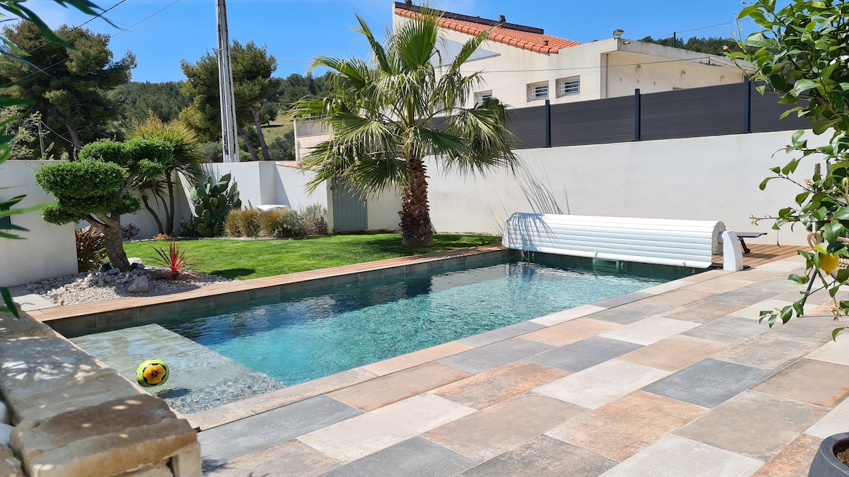 A heated swimming pool is framed by paved tiles and bordered by a neatly maintained lawn, featuring palm trees and decorative plants. A rolling cover for the pool is visible, adding functionality to the outdoor space.