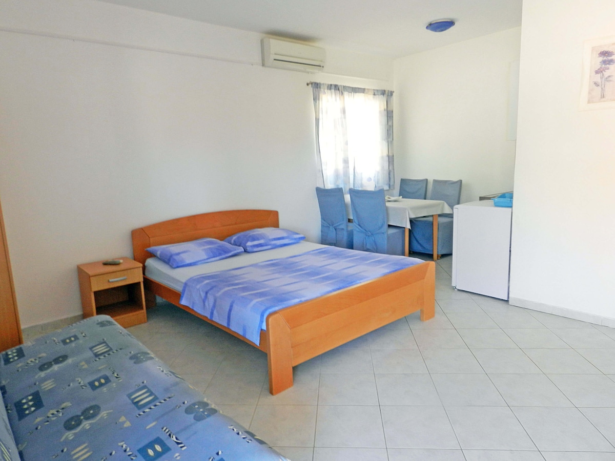 A compact studio apartment features a wooden bed with a light blue blanket and two additional single pillows. A dining area with blue chairs and a small table is visible, alongside a small fridge and an air conditioning unit. Natural light enters through a sheer window.