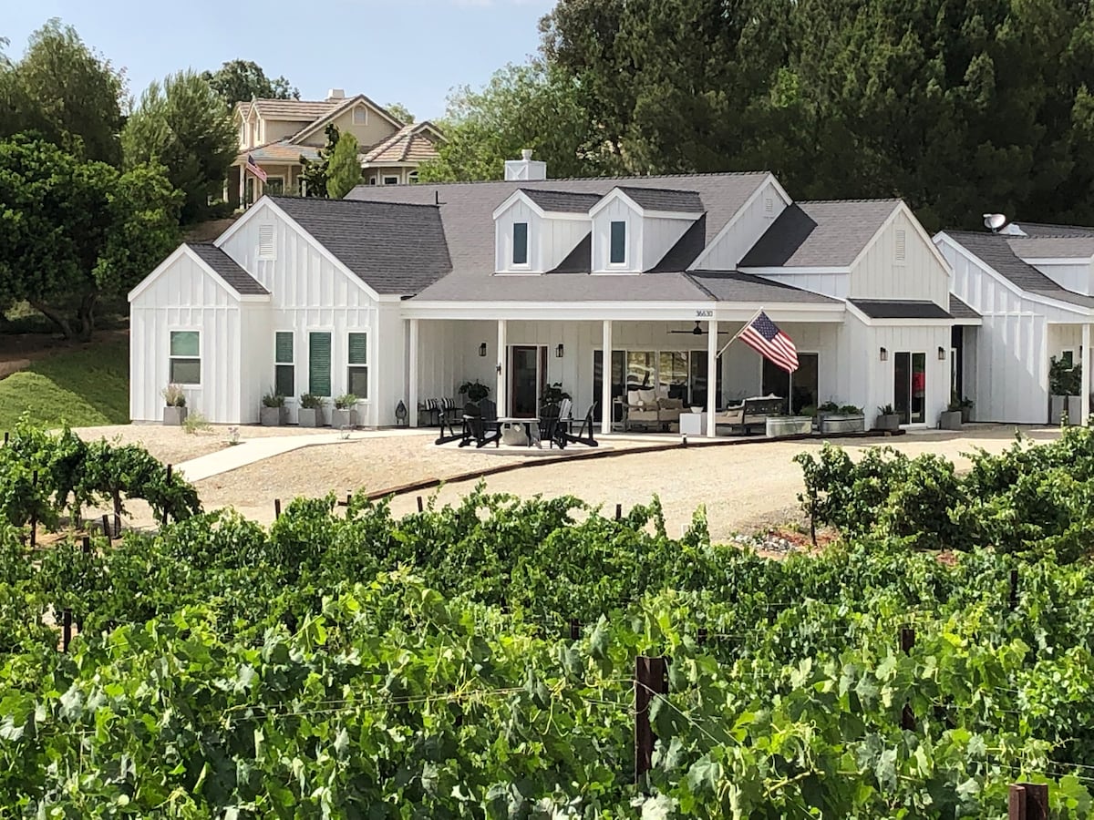 A modern farmhouse with a white exterior is set against a backdrop of lush vineyards. The entry features a spacious porch with black outdoor seating and an American flag. Surrounding grapevines are visible, adding to the serene Wine Country ambiance.