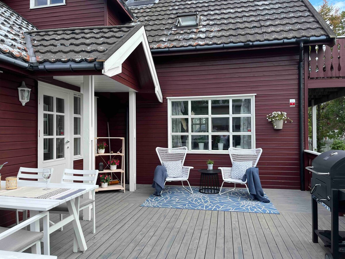 A charming terrace is depicted, featuring a wooden deck with a patio table and two white, woven chairs. A soft blue rug is placed underfoot, accompanied by a small potted plant. A grill is positioned nearby, enhancing the outdoor seating area.
