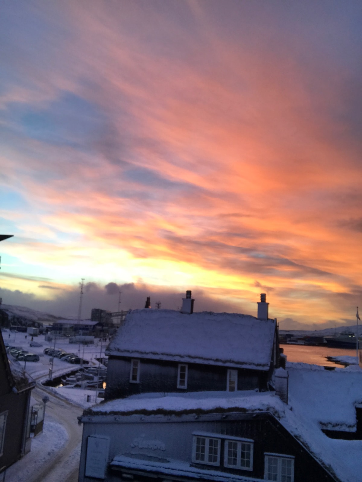 A vibrant sky is displayed at sunset, featuring shades of orange and pink. Snow-covered rooftops and a clear view of a harbor are visible in the foreground, with streets lined by parked vehicles that lead towards the water.