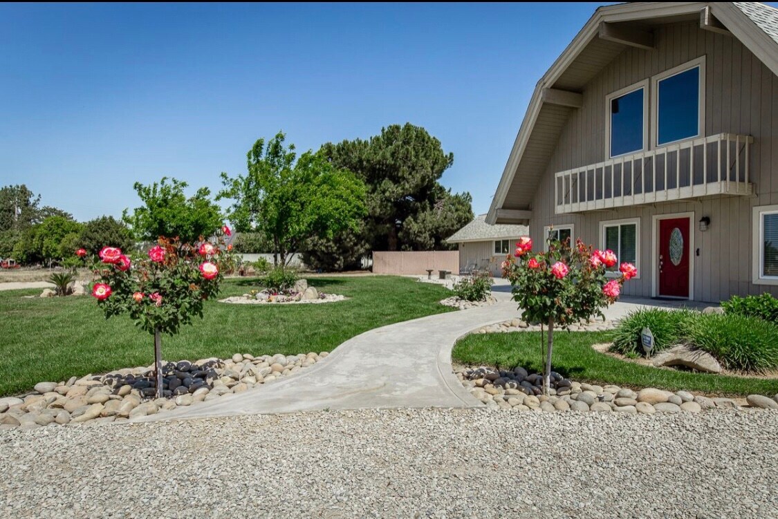 A welcoming front entrance is framed by neatly landscaped gardens featuring rose bushes. A winding path leads to a spacious two-story house with large windows and a red door, set against a backdrop of green trees and a clear blue sky.