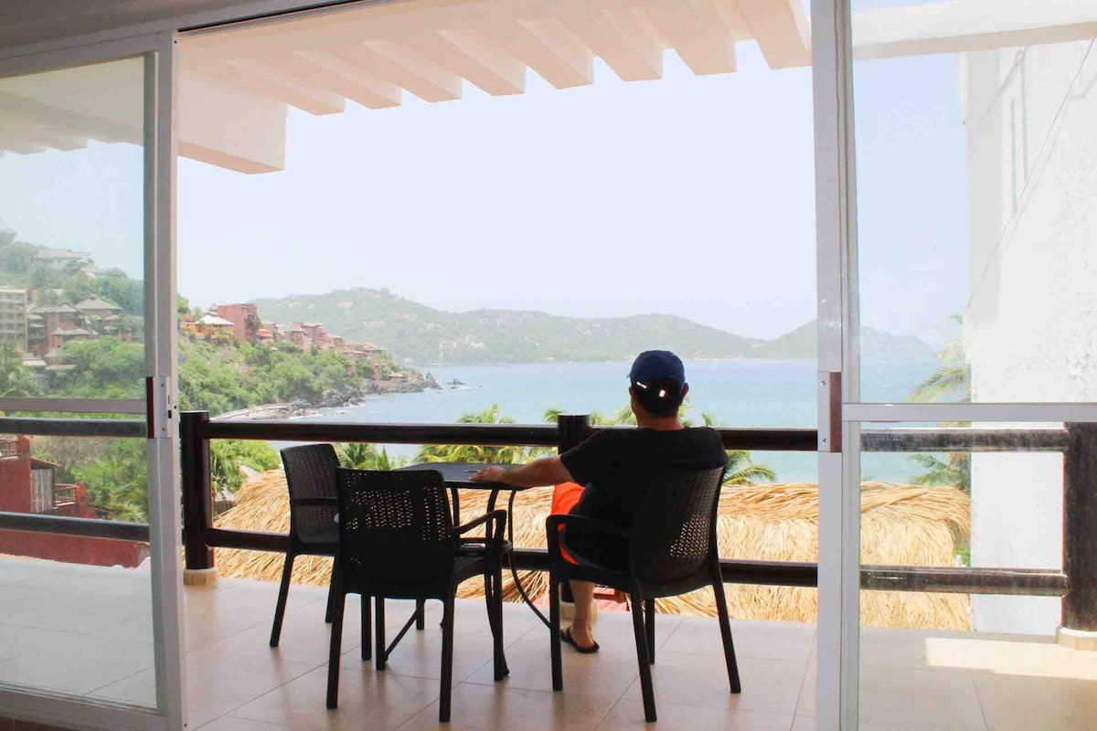 A spacious balcony with outdoor seating provides a stunning view of the bay. The area is surrounded by lush greenery and distant hills, creating a serene atmosphere for relaxation. Floor-to-ceiling glass doors bring in natural light, enhancing the connection with the outdoors.