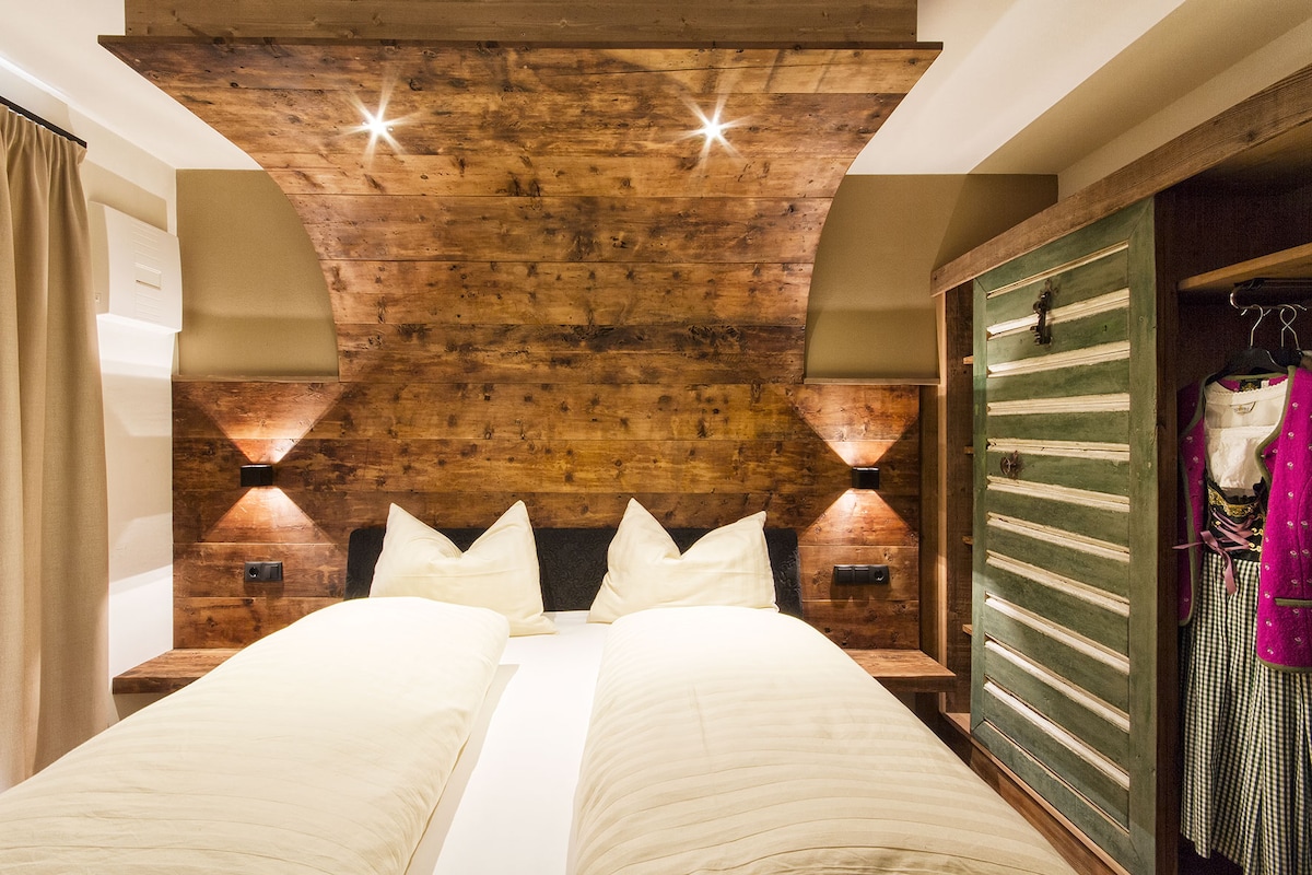 A cozy bedroom features a rustic wood-paneled headboard with integrated lighting. The spacious bed is adorned with light bedding, while contrasting storage spaces reflect a traditional design. A wardrobe with vibrant green accents and hanging space is visible, enhancing the room's overall warmth.