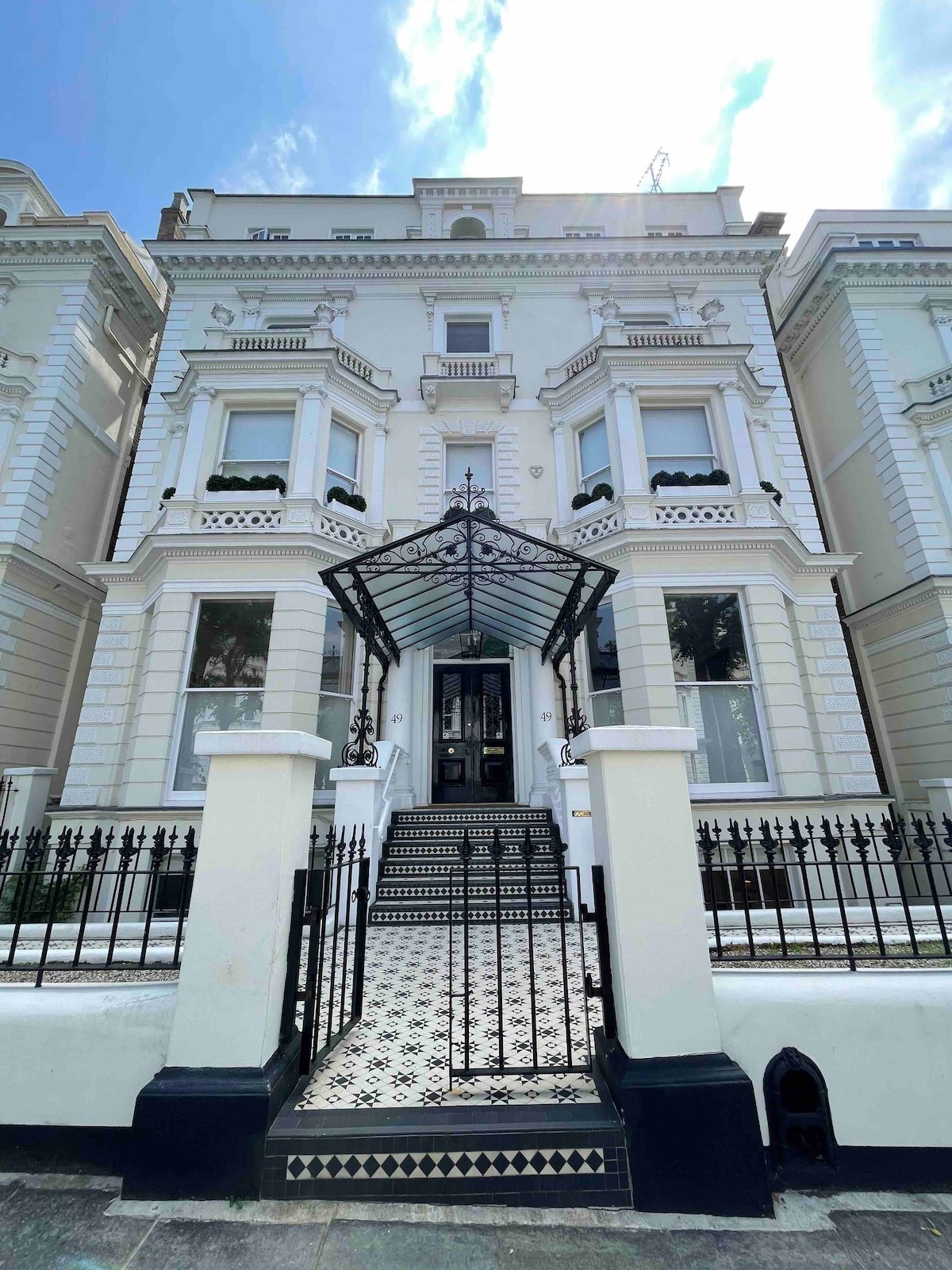 The elegant facade of the building is framed by intricate black wrought iron gates and a decorative canopy. A patterned tiled pathway leads to the entrance, which features large windows that allow natural light to fill the interior. The architecture showcases classic details, including ornate trim and a symmetrical design.