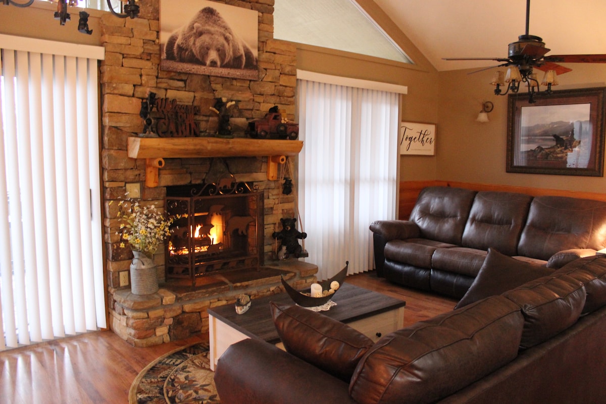 A spacious living room features a stone fireplace with a bear motif above it. Large windows with blinds allow natural light to fill the area, while a leather sectional sofa and a coffee table provide comfortable seating. Decorative accents include floral arrangements and wall art.