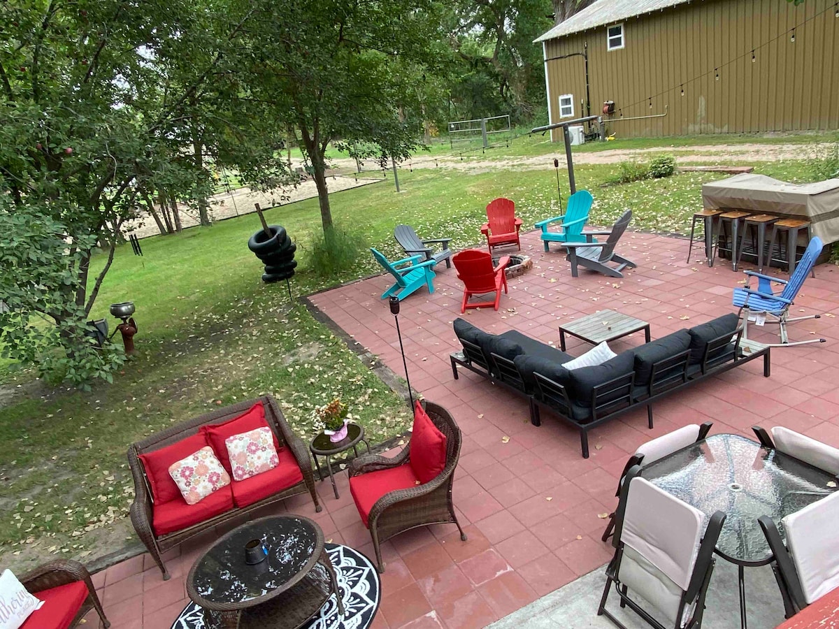 A spacious outdoor patio features various seating options, including lounge chairs and a sectional sofa arranged around a coffee table. Bright red and blue adirondack chairs complement wicker seating, and string lights hang overhead, creating a welcoming environment for relaxation and gatherings.