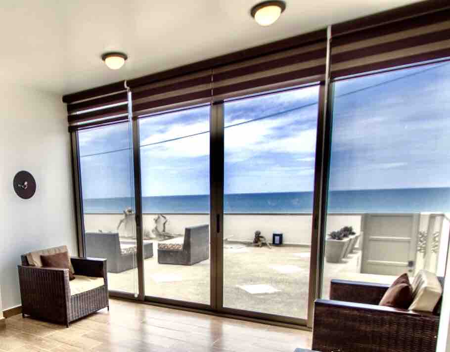 Large sliding glass doors provide a clear view of the ocean, allowing natural light to fill the space. Comfortable seating is arranged in a cozy configuration, with a neutral color palette complementing the serene atmosphere of the room.